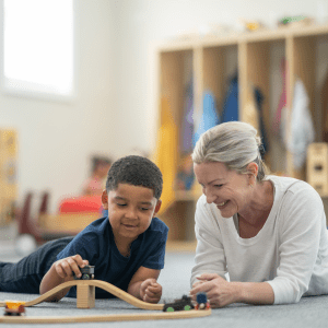 A young boy and a Pioneer travel therapy specialist playing with a wooden train set on the floor. 