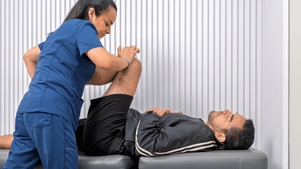A female sports physical therapist in blue scrubs assisting a male patient with hip and leg stretches during a rehabilitation session.