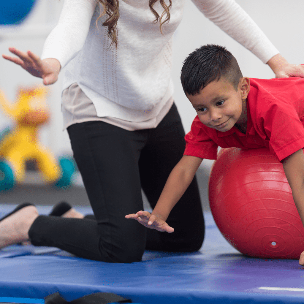 Pediatric physical therapist sitting on floor mat with smiling young boy balanced on red exercise ball during therapy session.